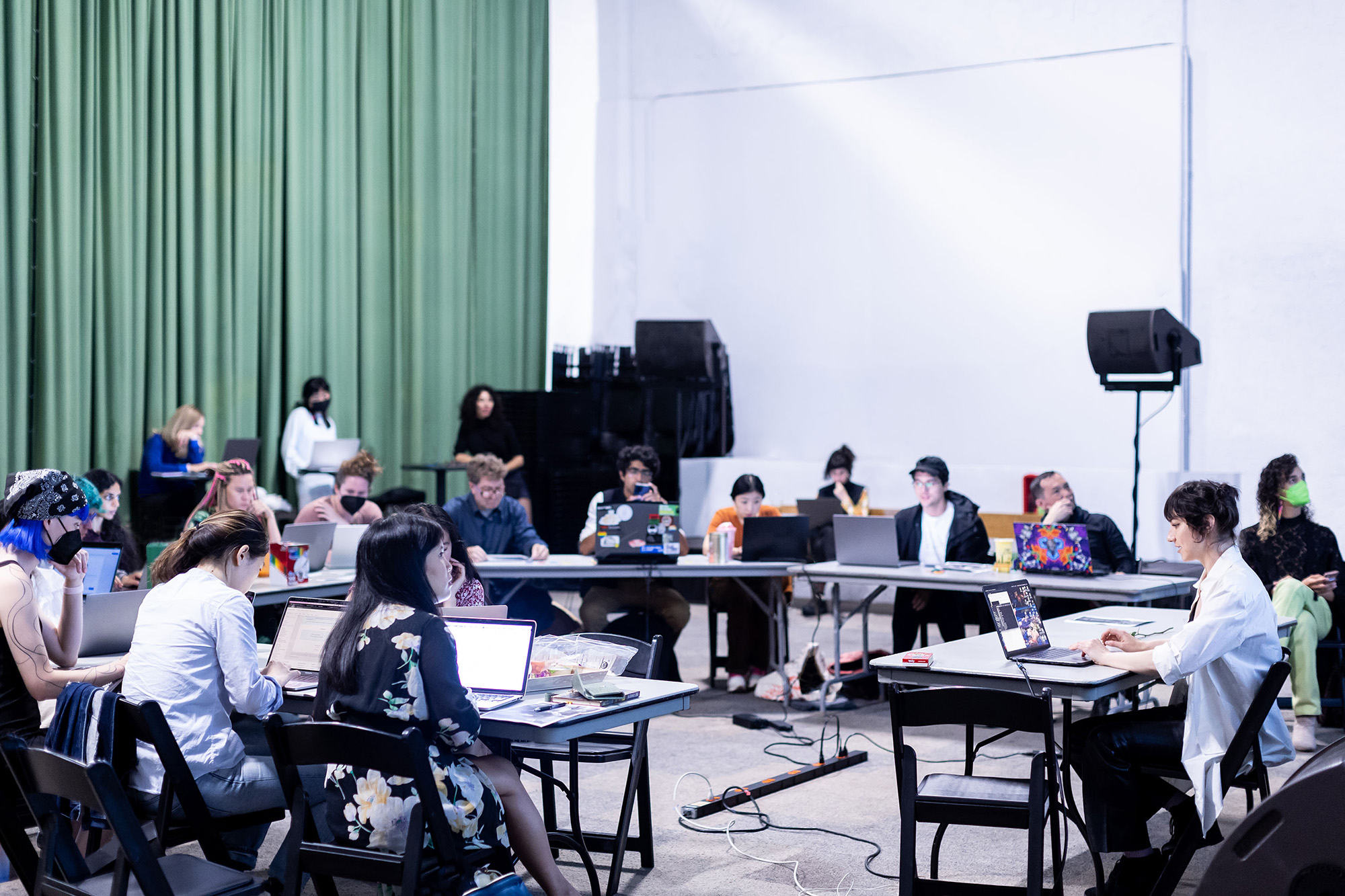 15 people sit around a circle of tables, with laptops and art supplies strewn around them.