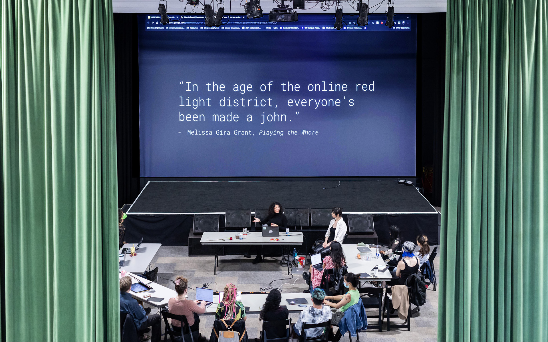 Workshop participants sit gathered before a large screen, where a slide displays a quote by the author Melissa Gira Grant: 