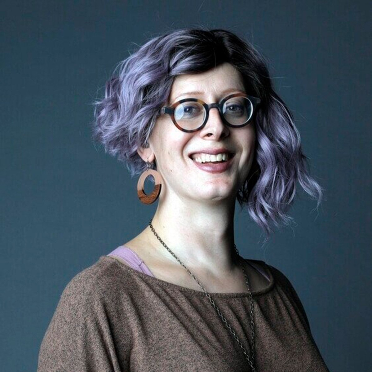 In a smiling photo of Sasha Costanza-Chock, her wavy, purple-toned hair and bold earrings stand out from a deep blue background.