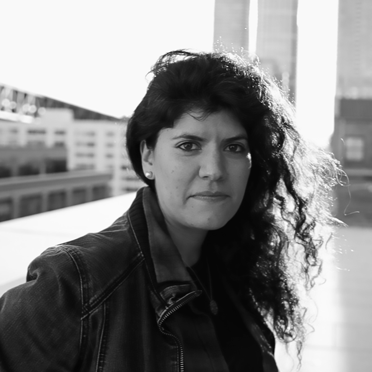 In a black and white photo, Joana Varon poses in front of a cityscape, looking intently at the viewer.