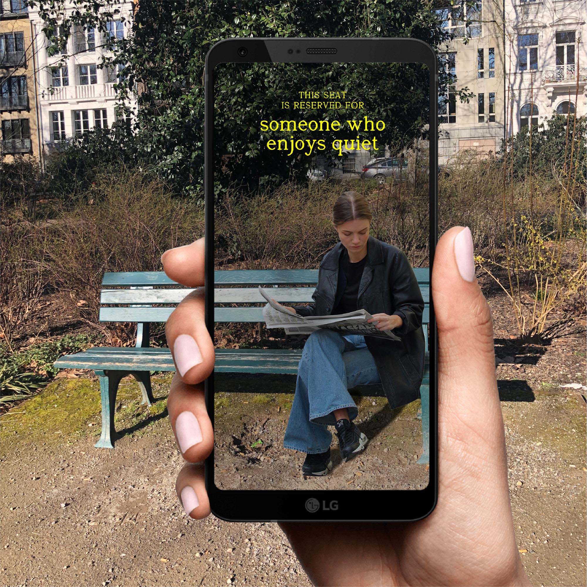 A woman sits reading a newspaper on a green park bench on a sunny day, framed by a hand holding up a smartphone whose screen reads, 