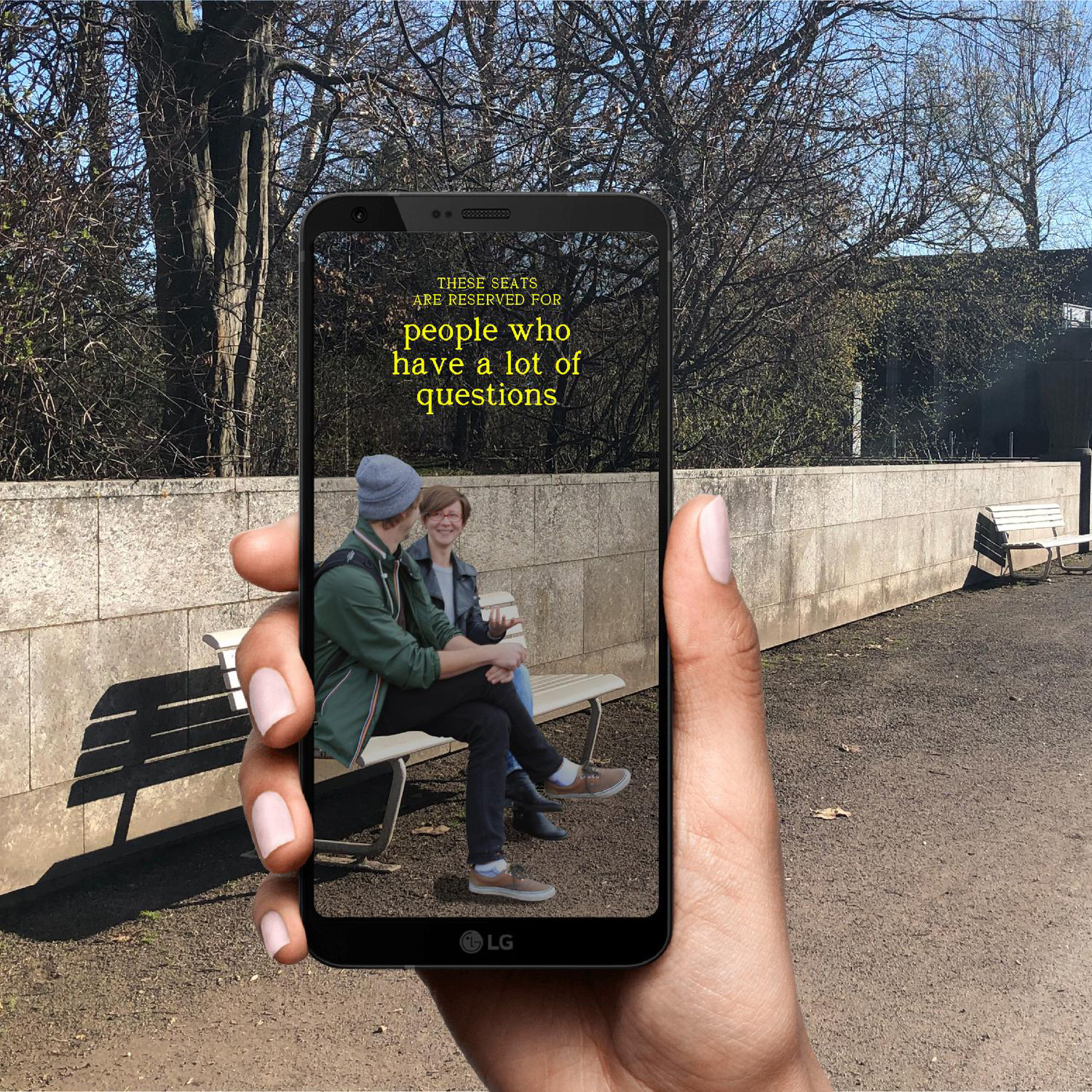 Two people chatting on a park bench on a sunny day are framed by a hand holding up a smartphone whose screen reads, 