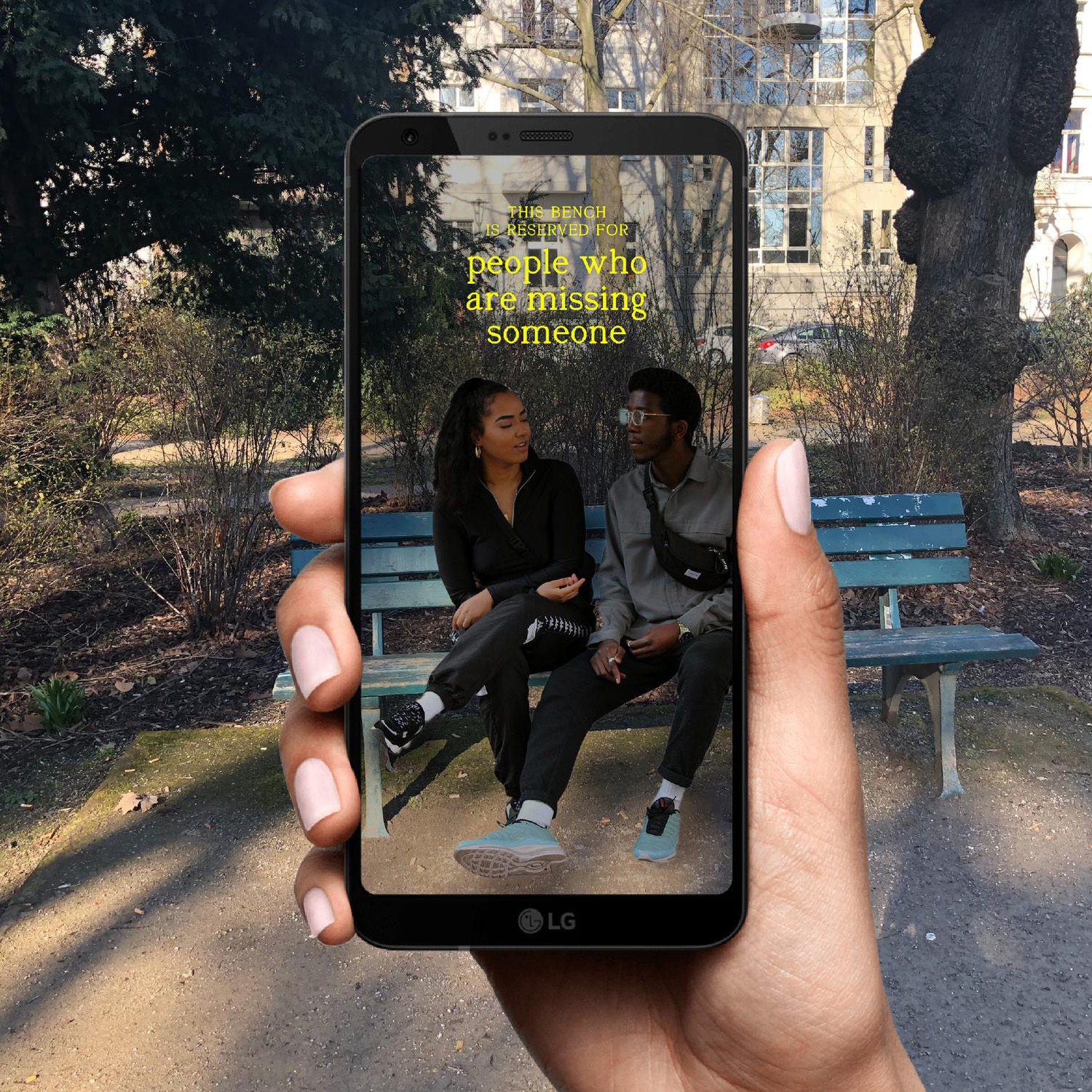 Two people chatting on a park bench on a sunny day are framed by a hand holding up a smartphone whose screen reads, 