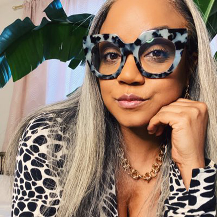 A photo of Sinnamon Love shows a Black woman with chunky, tortoise shell glasses frames that echo a bold, graphic shirt.