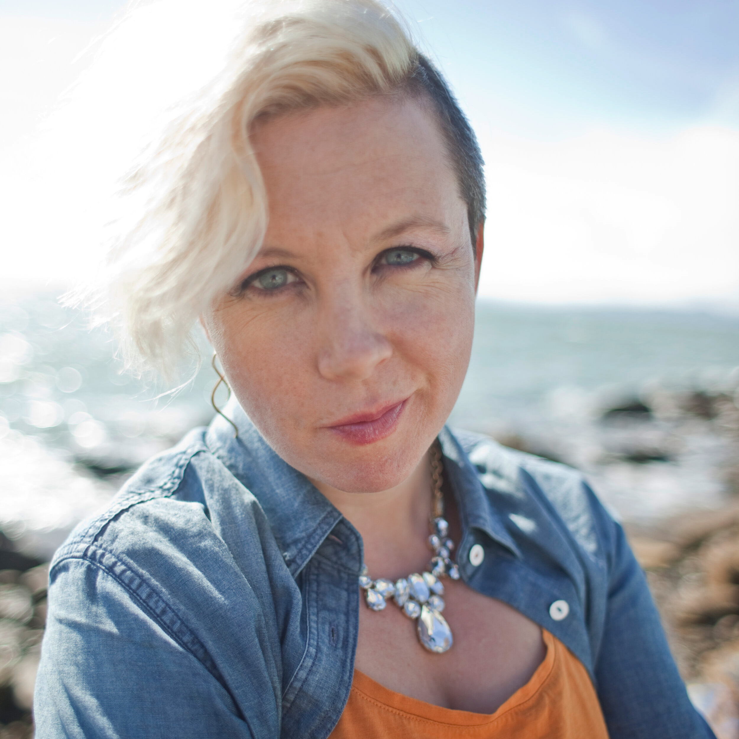 A photo of Tina Horn shows a woman on the beach, her blue eyes echoed in a denim shirt and the ocean behind her.